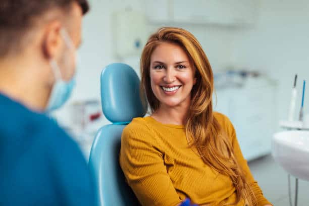 A happy woman with long red hair smiling at the camera while sitting in a dental chair, with a blurred male dentist wearing a mask in the foreground, suggesting a positive dental visit or successful treatment