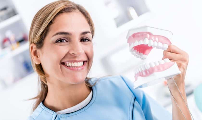 A smiling female dentist or dental assistant, wearing light blue scrubs, is holding a large model of a set of dentures.