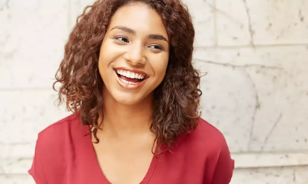 A medium shot of a young woman with curly brown hair, wearing a red top. She is smiling after successful cosmetic dental treatment broadly and looking off to the side.
