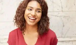 A medium shot of a young woman with curly brown hair, wearing a red top. She is smiling after successful cosmetic dental treatment broadly and looking off to the side.