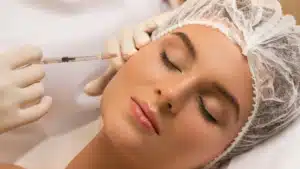 A close-up of a woman lying down with her eyes closed, wearing a hair net, receiving a cosmetic injection near her temple from a professional wearing white gloves and holding a syringe.